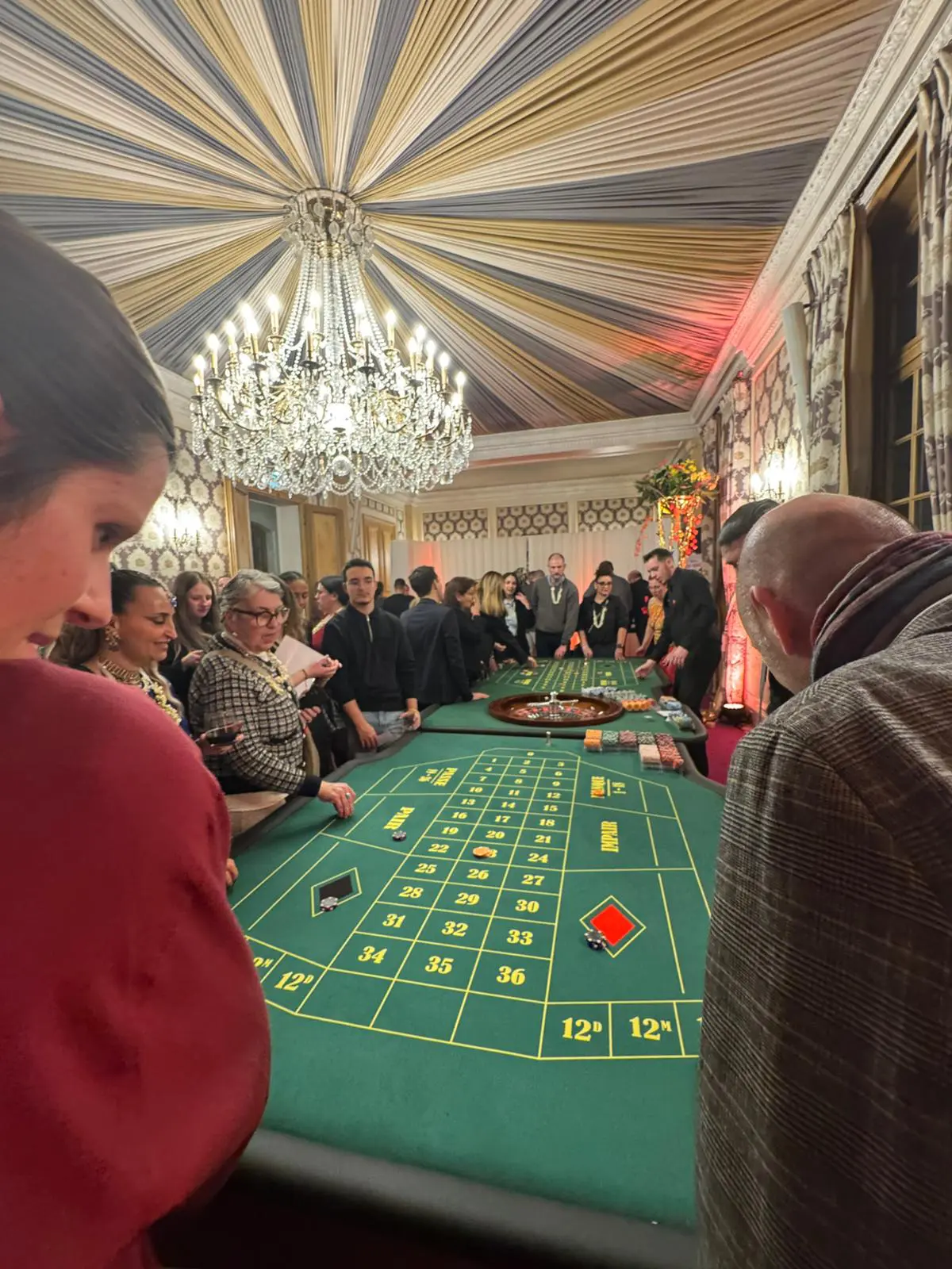 Ambiance immersive d'une soirée casino factice à Paris : grande table de roulette entourée d'invités élégants, lustre cristal et plafond drapé dans un salon prestigieux – Animation professionnelle par L'As du Casino.